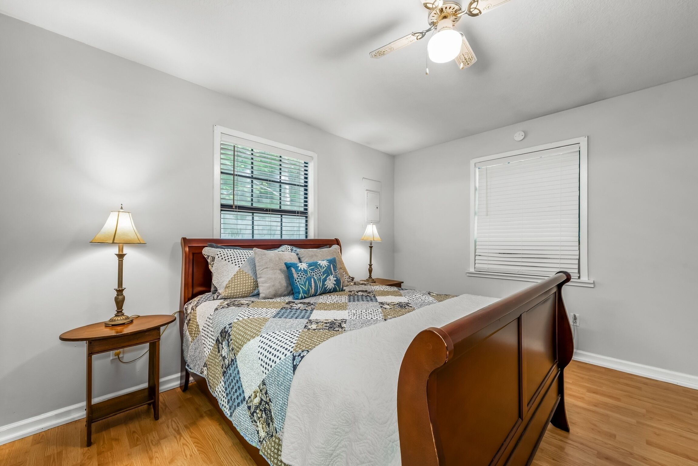 A bedroom with a wooden bed, two bedside lamps, a window with blinds, and a ceiling fan.
