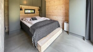 Une chambre moderne avec une tête de lit en bois, un lit recouvert d’une couverture grise, une petite fenêtre et un radiateur placé près du lit.