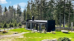 Une cabane moderne à la façade vitrée, entourée d’une forêt et d’une pelouse bien entretenue parsemée de rochers.