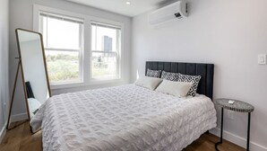 A bedroom with a large bed, a standing mirror, a small table, and an air conditioning unit. - 3BR2Bth Parking, Cambridge/Boston, W/D, Subway,A/C (Somerville)
