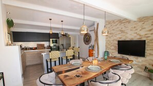 Una cocina moderna con armarios oscuros, microondas y refrigerador. Hay un comedor con mesa de madera, sillas y un televisor montado en la pared.