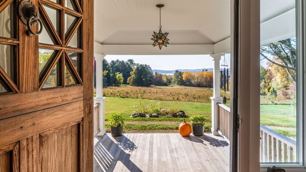 Un portico in legno con una lampada a sospensione a forma di stella, piante in vaso e vista su un campo erboso con alberi e montagne all'orizzonte.