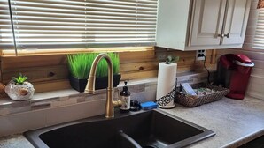 A kitchen with a black sink, white cabinets, a window with blinds, a coffee maker, and a small plant.