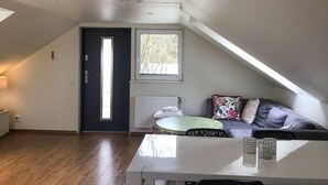 A room with a sloped ceiling, a door, a window, a radiator, a sofa with cushions, a table, and a lamp. - 2 bedroom gorgeous home in åkersberga (Åkersberga)