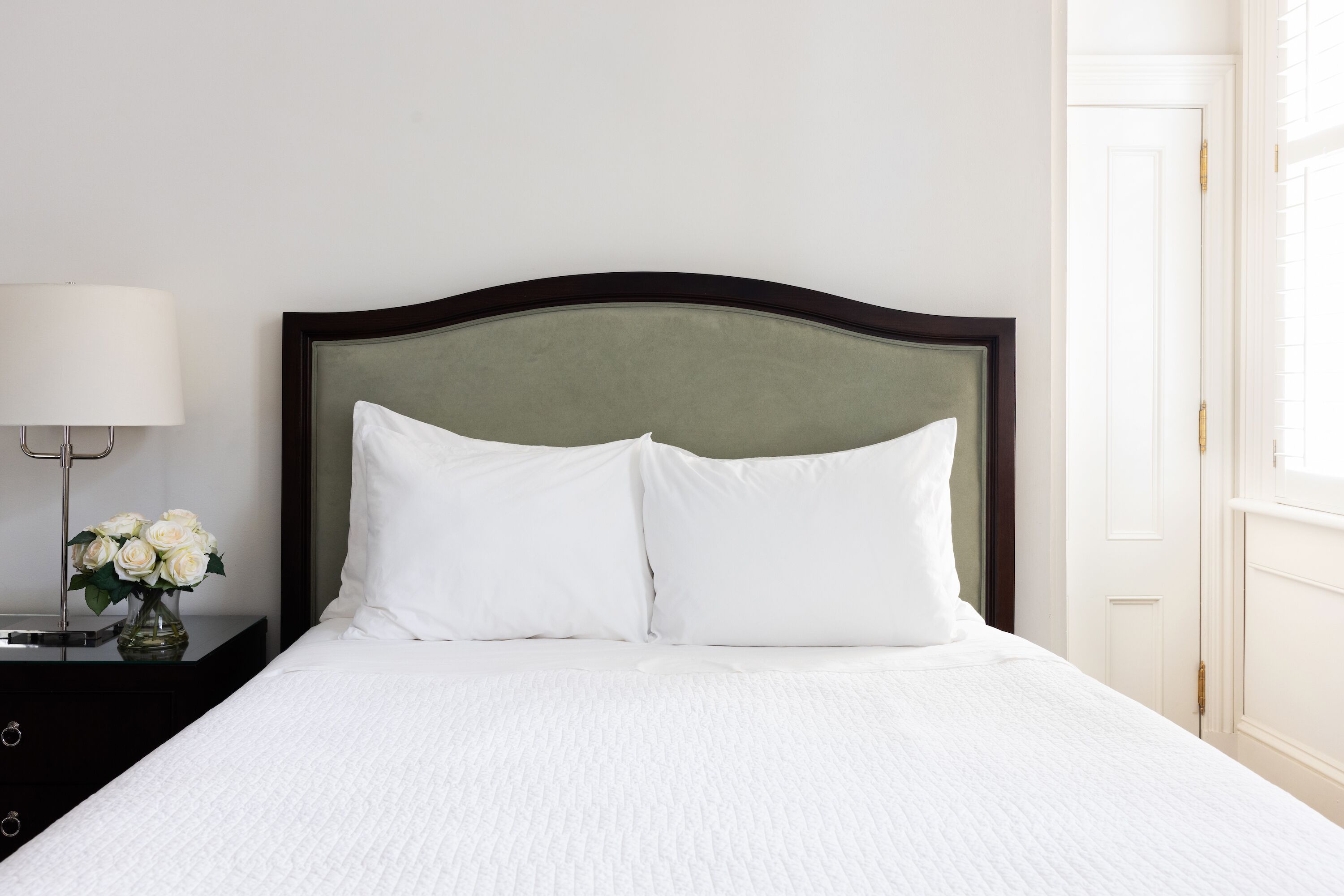 Un letto rifatto con lenzuola bianche e una testiera scura, un comodino con una lampada e un vaso di fiori, e una porta bianca sullo sfondo.