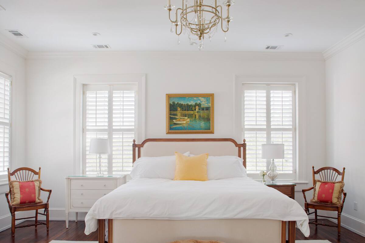 Quarto com cama grande, um banquinho estofado, duas cadeiras de madeira, mesa de cabeceira, abajur e um quadro na parede.