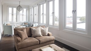 Een ruime woonkamer met een beige bank, een lichtblauwe fauteuil en een witte salontafel. Grote ramen zorgen voor veel daglicht en uitzicht op het omliggende landschap.