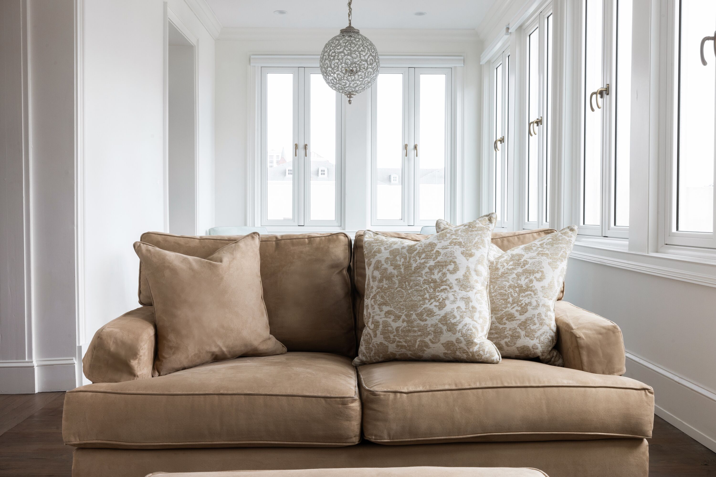 Un canapé d’angle beige agrémenté de coussins à motifs, dans une pièce lumineuse aux murs blancs, dotée de grandes fenêtres et d’un luminaire suspendu.