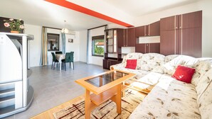 Ein modernes Wohnzimmer mit einer Couch, einem Couchtisch und einem Essbereich mit Stühlen. Es gibt eine Küchenzeile inklusive Kühlschrank und ein Fenster.