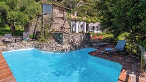 Une piscine aux murs en pierre et au bord en bois, entourée de verdure et d’un petit bâtiment.