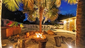 A nighttime outdoor area with a fire pit surrounded by wooden stumps, palm trees, and a swimming pool.