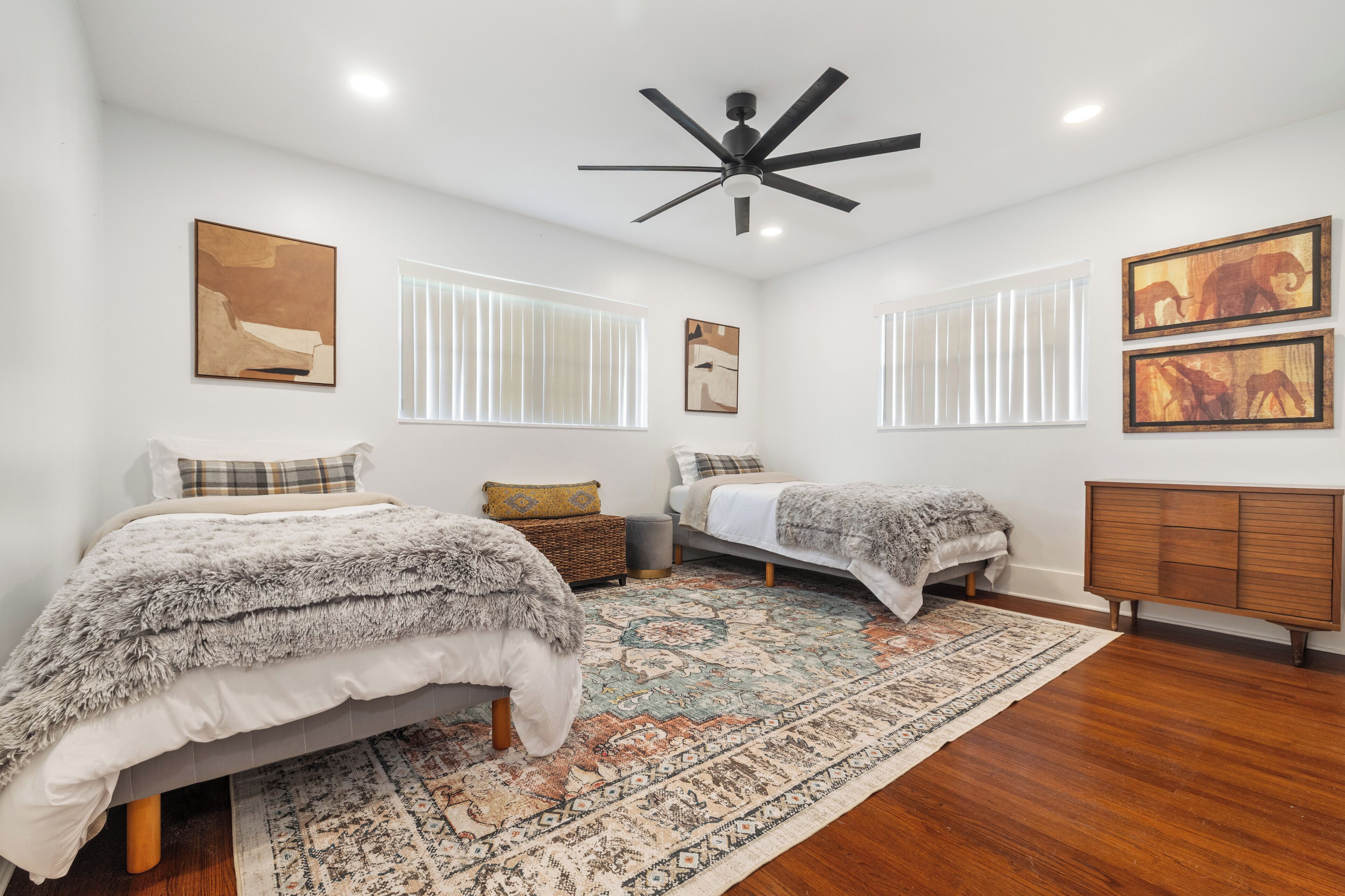 A bedroom with two beds, a ceiling fan, a large rug, and artwork on the walls.