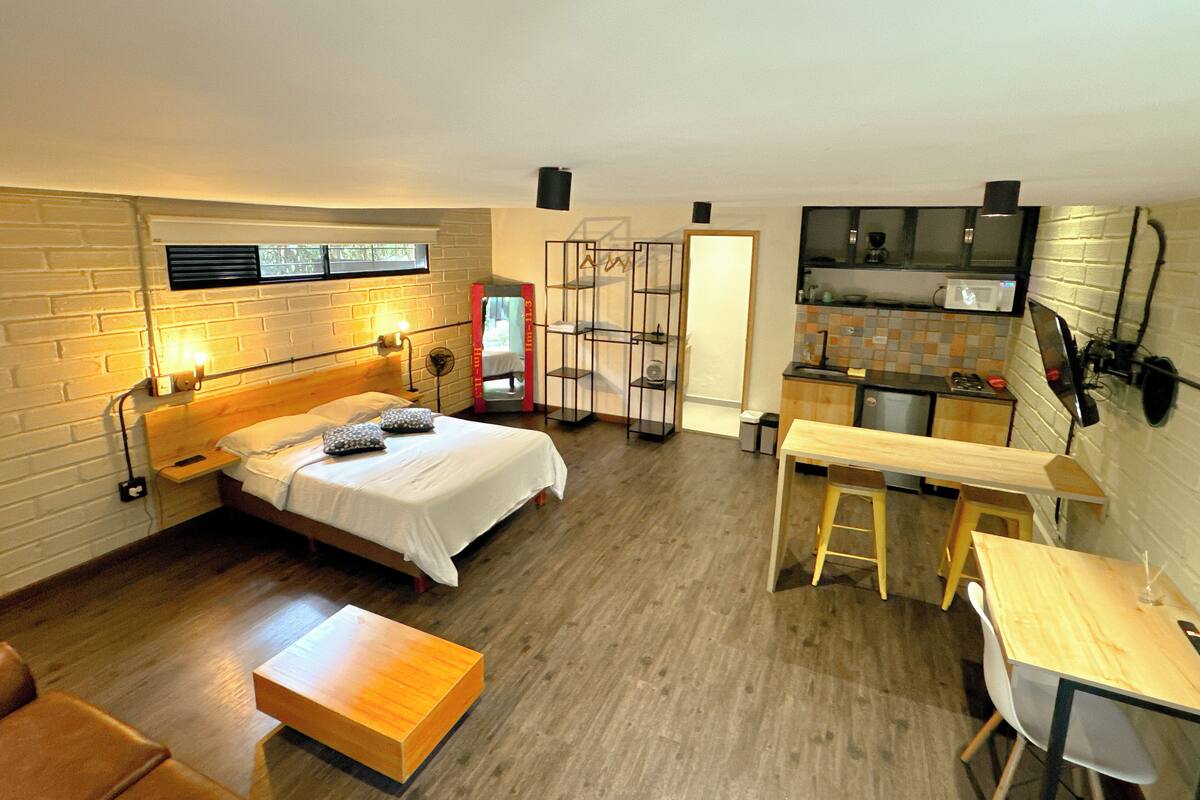Una habitación pequeña con cama, una mesita, un banco y un rincón de cocina.