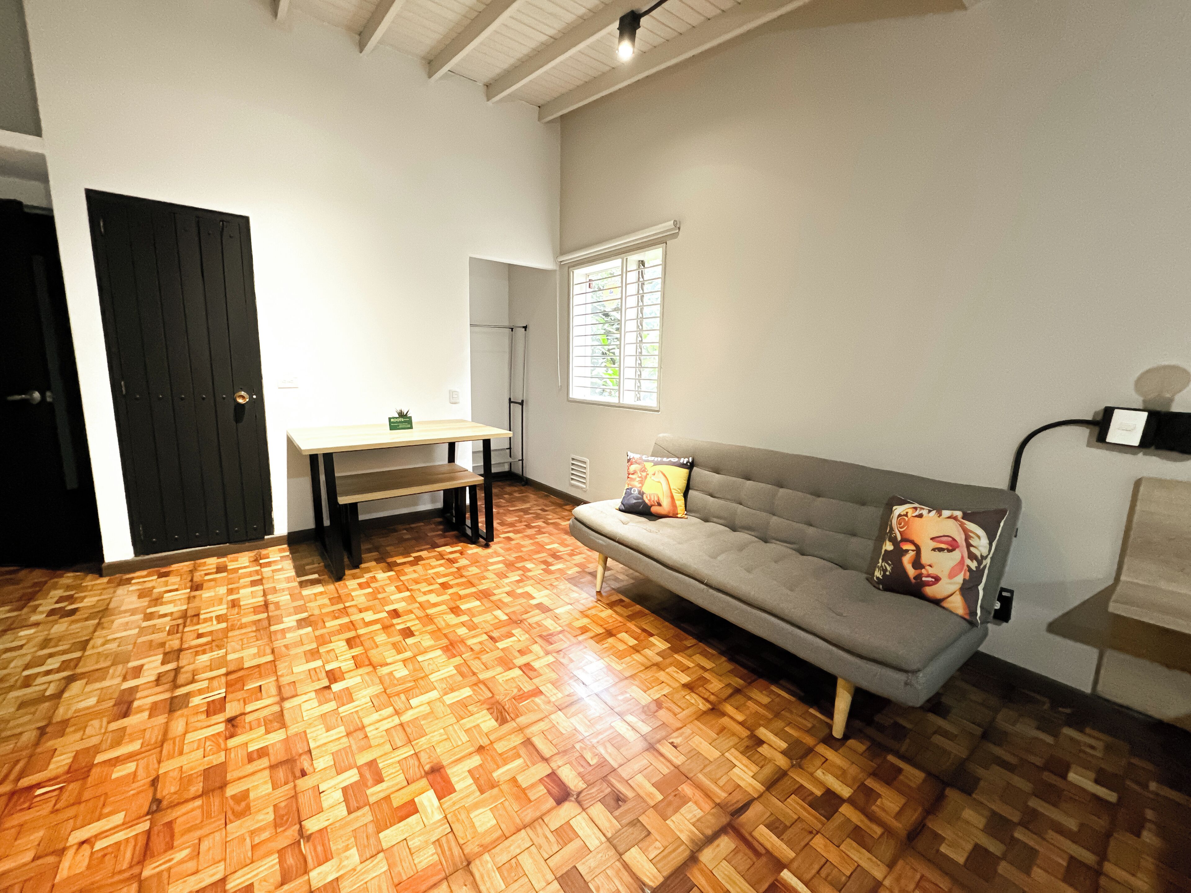 A room with a grey sofa, a wooden floor, a small table, a black door, and a window with blinds.