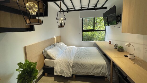 A compact bedroom with a bed, bedside table, and a window with a view of greenery. - Frontera by Roots Rentals (Medellín)