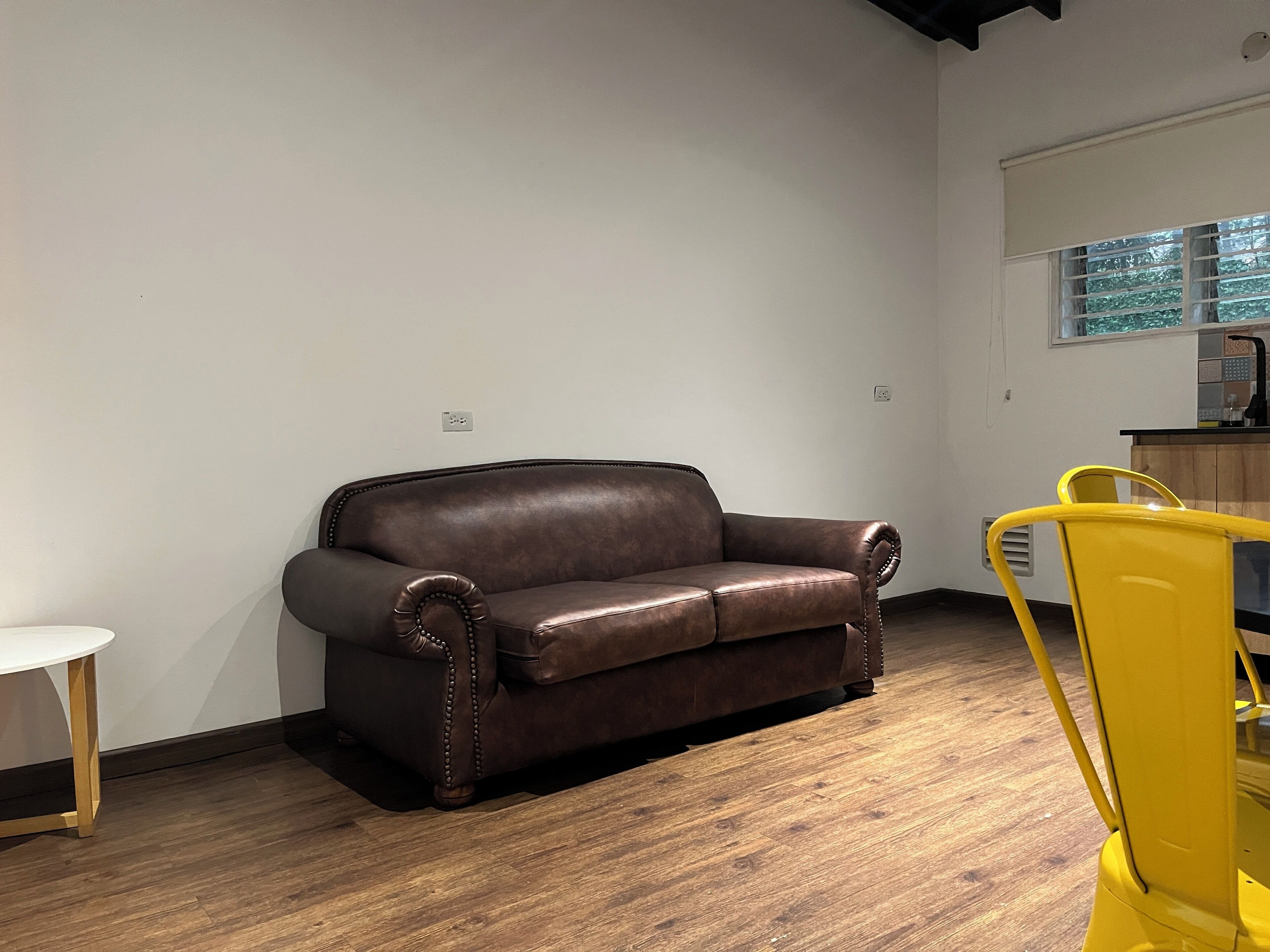 A room with a brown leather sofa, a yellow chair, a small white table, and a window with blinds.