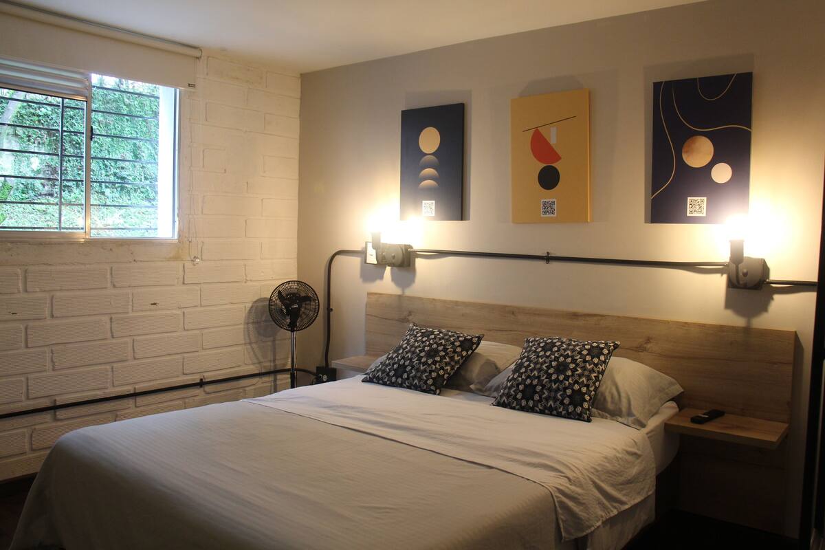 Una habitación de hotel con una cama, una mesita de noche, un ventilador de pared y una obra de arte en la pared.