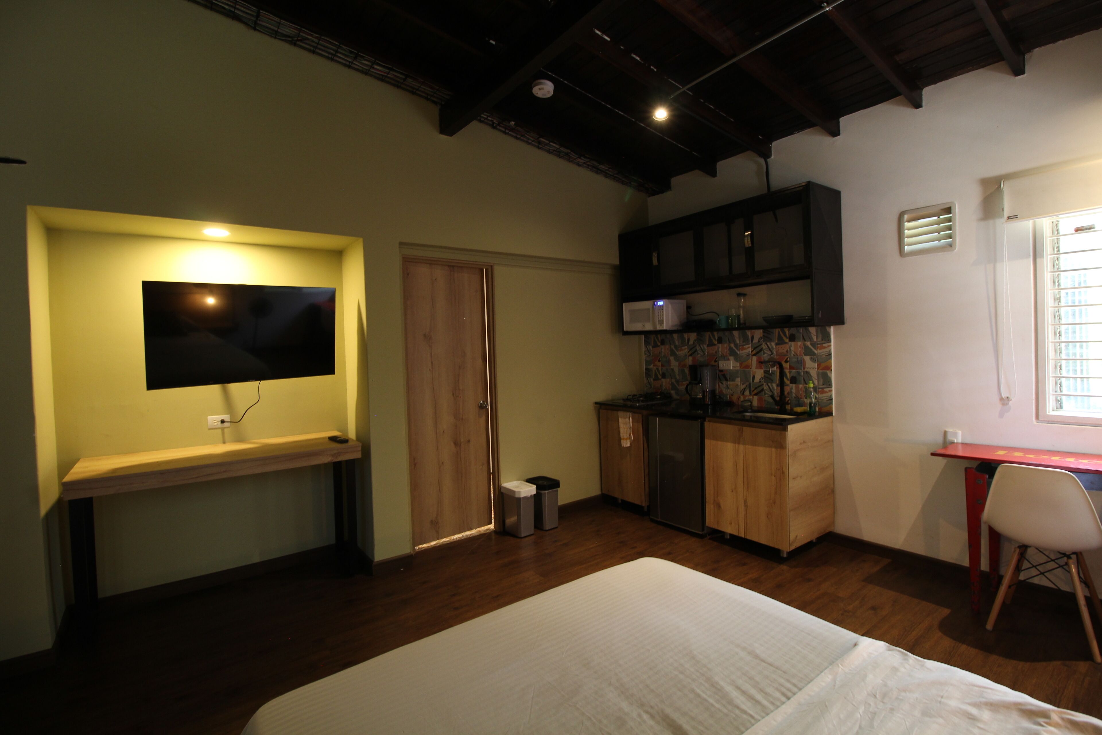 Une chambre de taille modeste comprenant un lit, un bureau, un coin cuisine et un téléviseur fixé au mur.