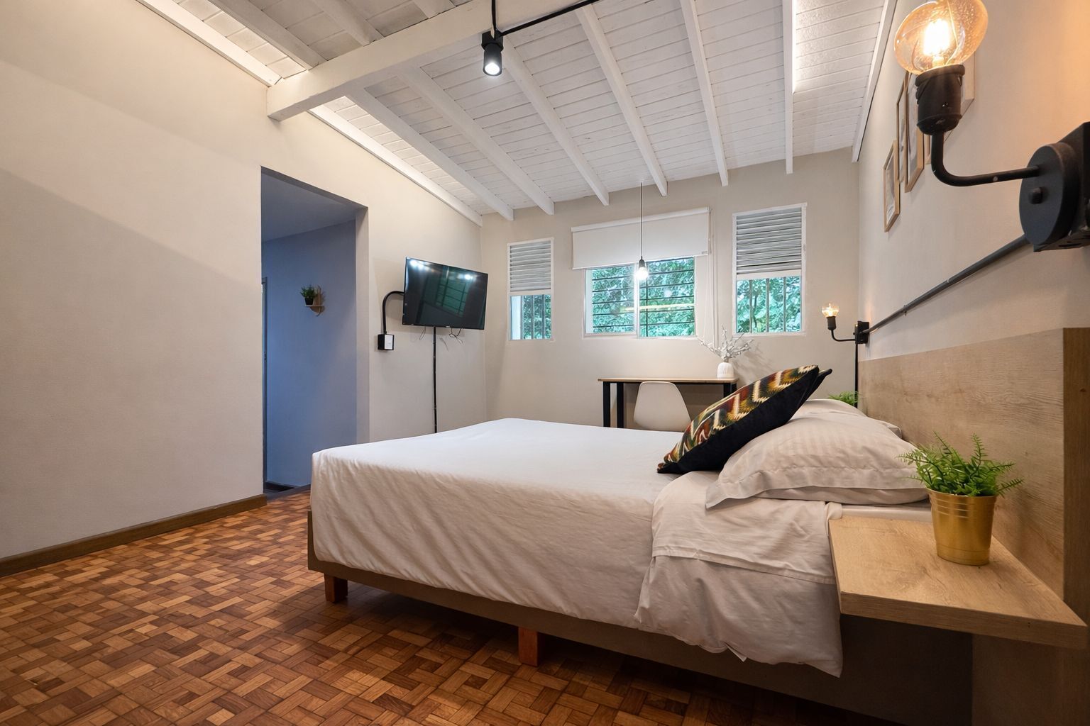 A bedroom with a bed, a desk, a TV, and a window with blinds.