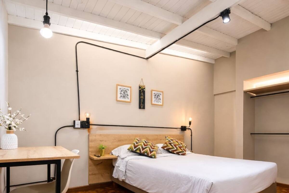 Un dormitorio con piso de madera, una cama con ropa blanca, una mesita de noche con lámpara y un estante fijado a la pared.