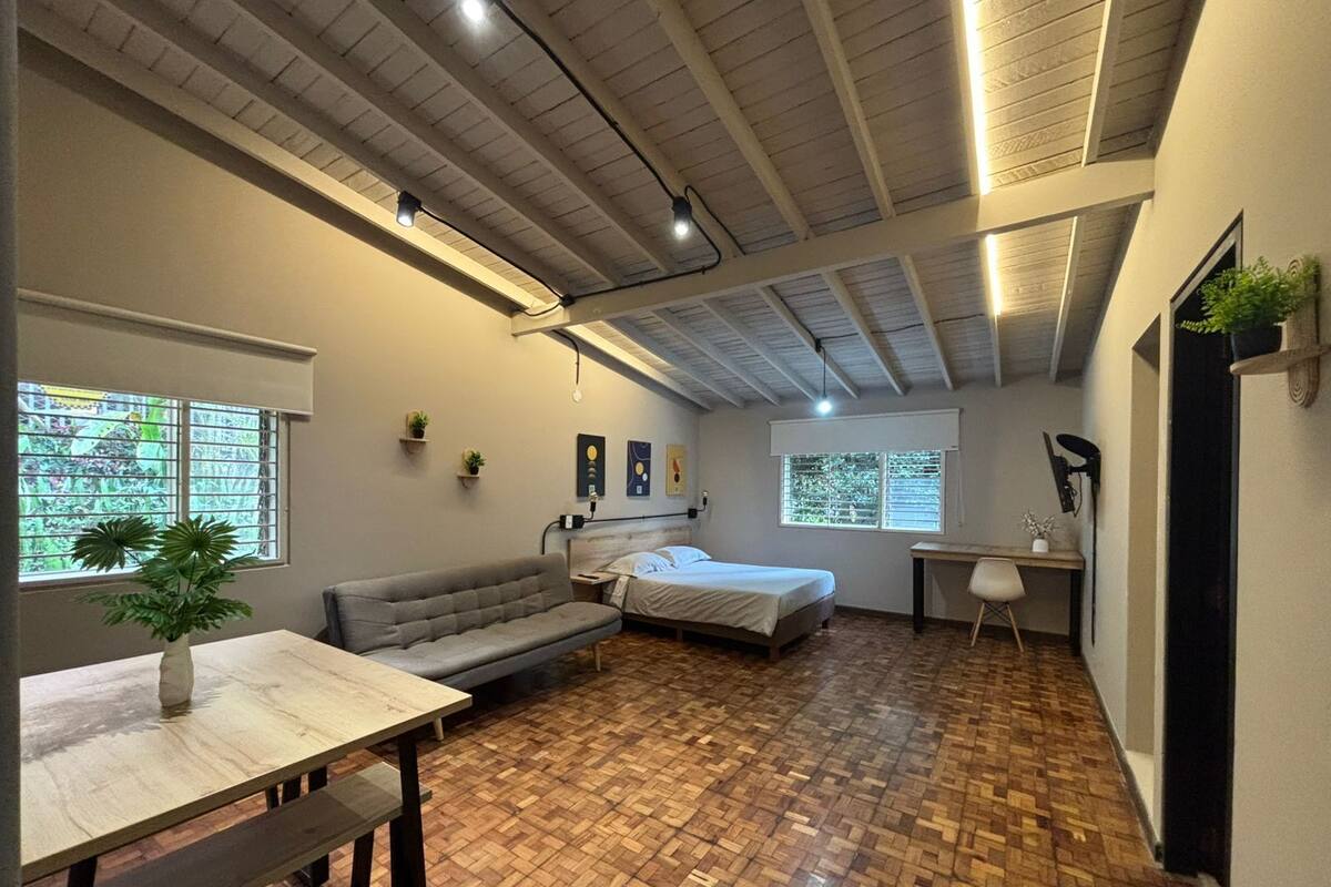 Una habitación con cama, sofá, escritorio y mesa. Hay dos ventanas con persianas, un techo con vigas a la vista y una planta sobre la mesa.