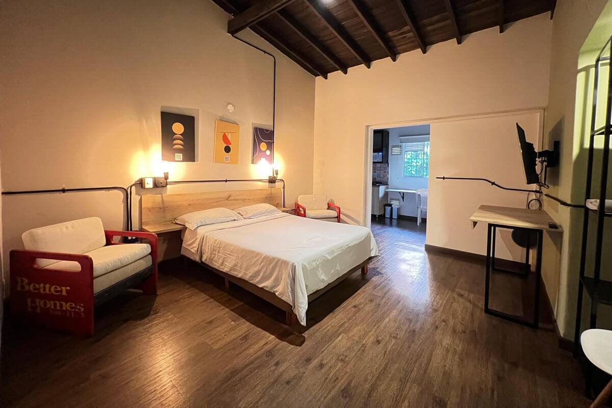 Un dormitorio con cama, mesitas de noche, una silla y un escritorio. Hay luces fijadas en la pared y obras de arte.