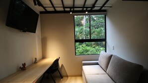 A room with a wooden desk, a grey sofa, a TV mounted on the wall, and a window with a view of greenery. - Frontera by Roots Rentals (Medellín)