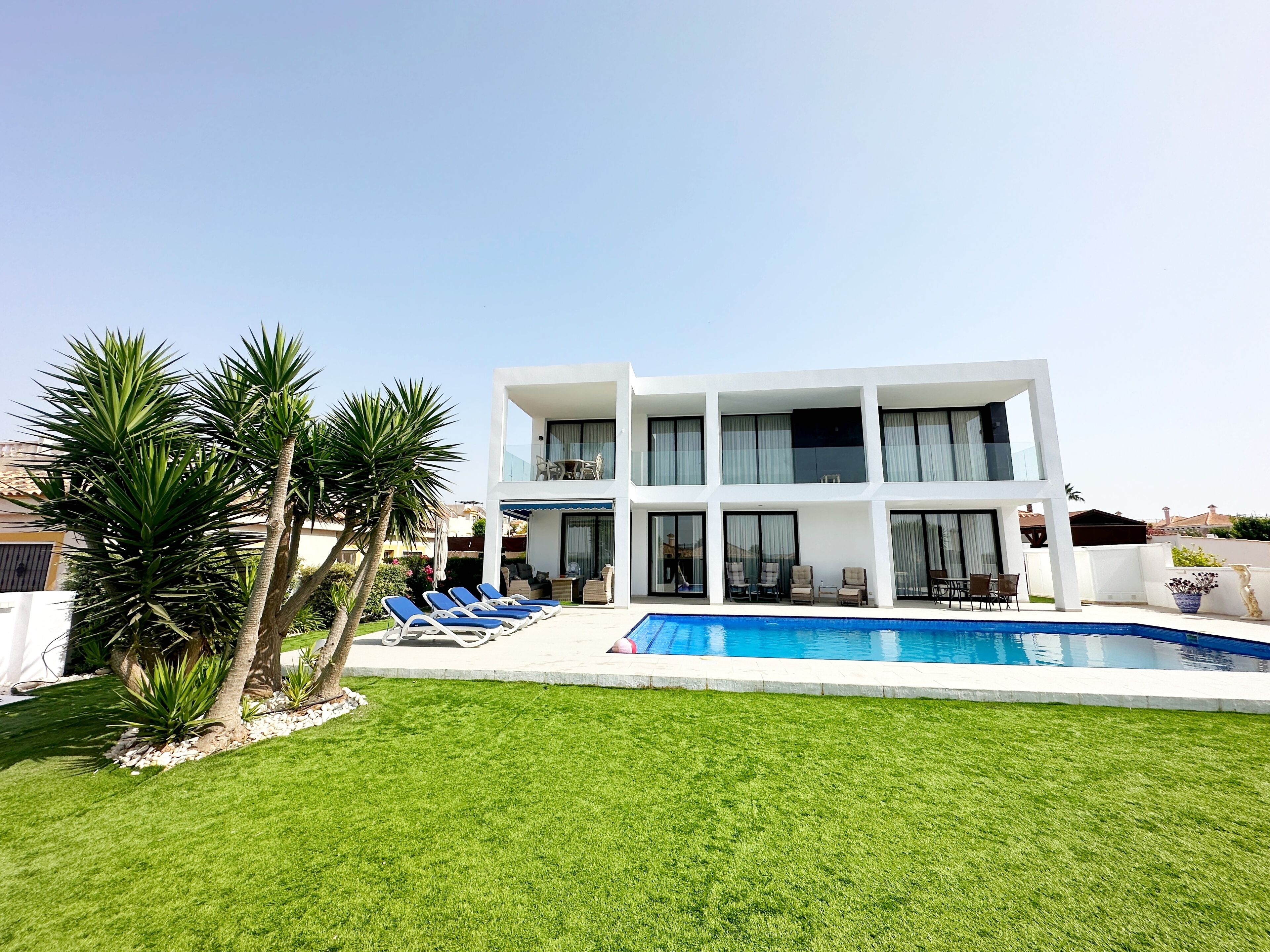 A modern two-story building with a swimming pool, sun loungers, and palm trees.