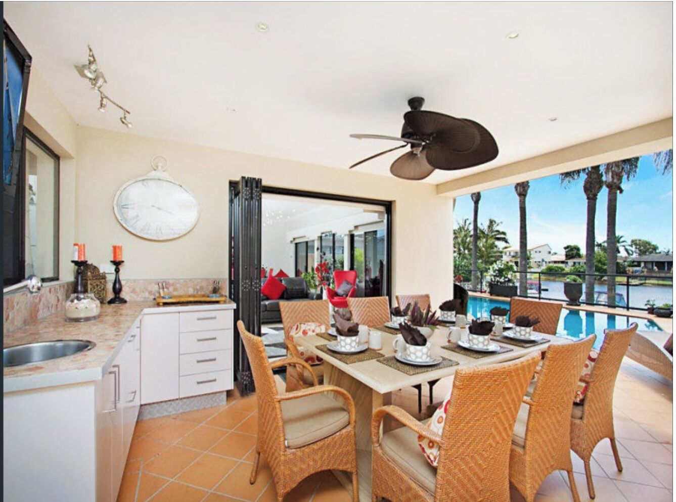 A dining area with a view of a pool and palm trees, featuring wicker chairs, a round table, and a ceiling fan.