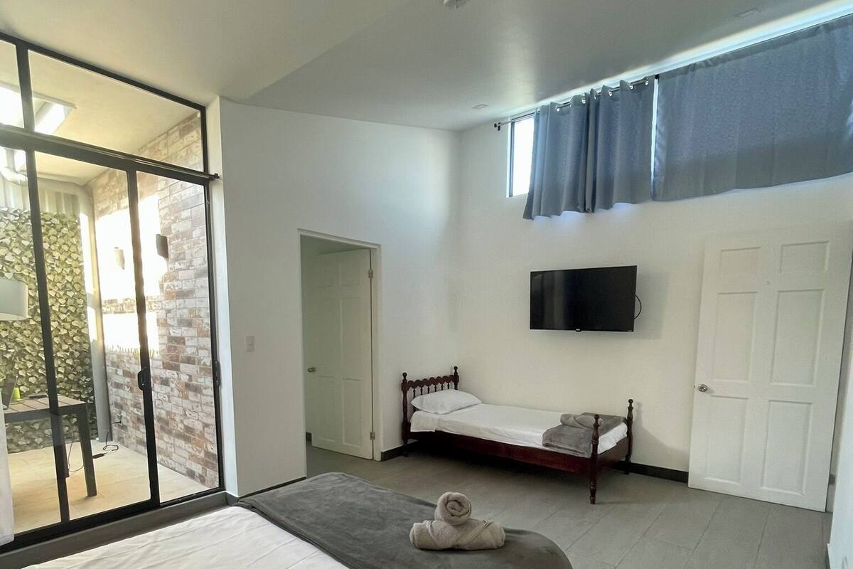 A bedroom with a large bed, a smaller bed, a TV mounted on the wall, and a sliding glass door leading to an outdoor area.