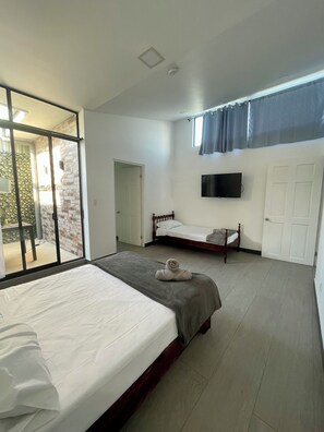 A bedroom with a large bed, a smaller bed, a TV mounted on the wall, and a sliding glass door leading to an outdoor area. - Casa Bellavista (San José)