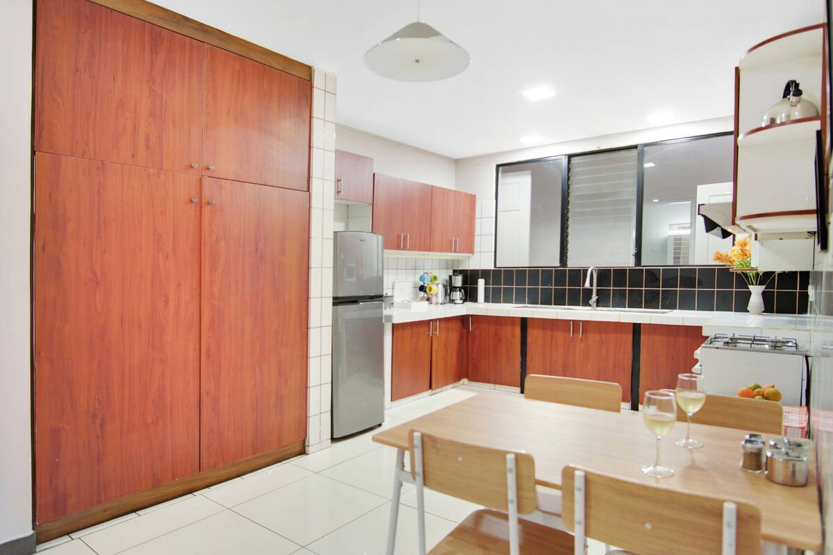 A modern kitchen with wooden cabinets, a stainless steel refrigerator, a dining table with chairs, and a vase with flowers on the counter.
