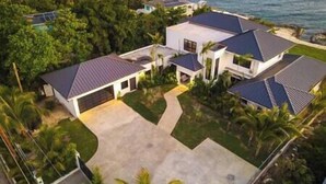 A modern two-story house with a covered driveway and a large front lawn, surrounded by lush greenery and a view of the ocean. - Ripple Negril, The ultimate escape. Unpack, Relax, Enjoy, Ripple Negril, The ultimate escape (West End)