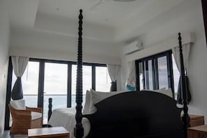 Ein Raum mit einem Himmelbett, großen Fenstern, Meerblick und Deckenventilator.
