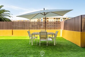 En terrasse med et bord og stole under en stor parasol, omgivet af et træhegn og en gul mur.