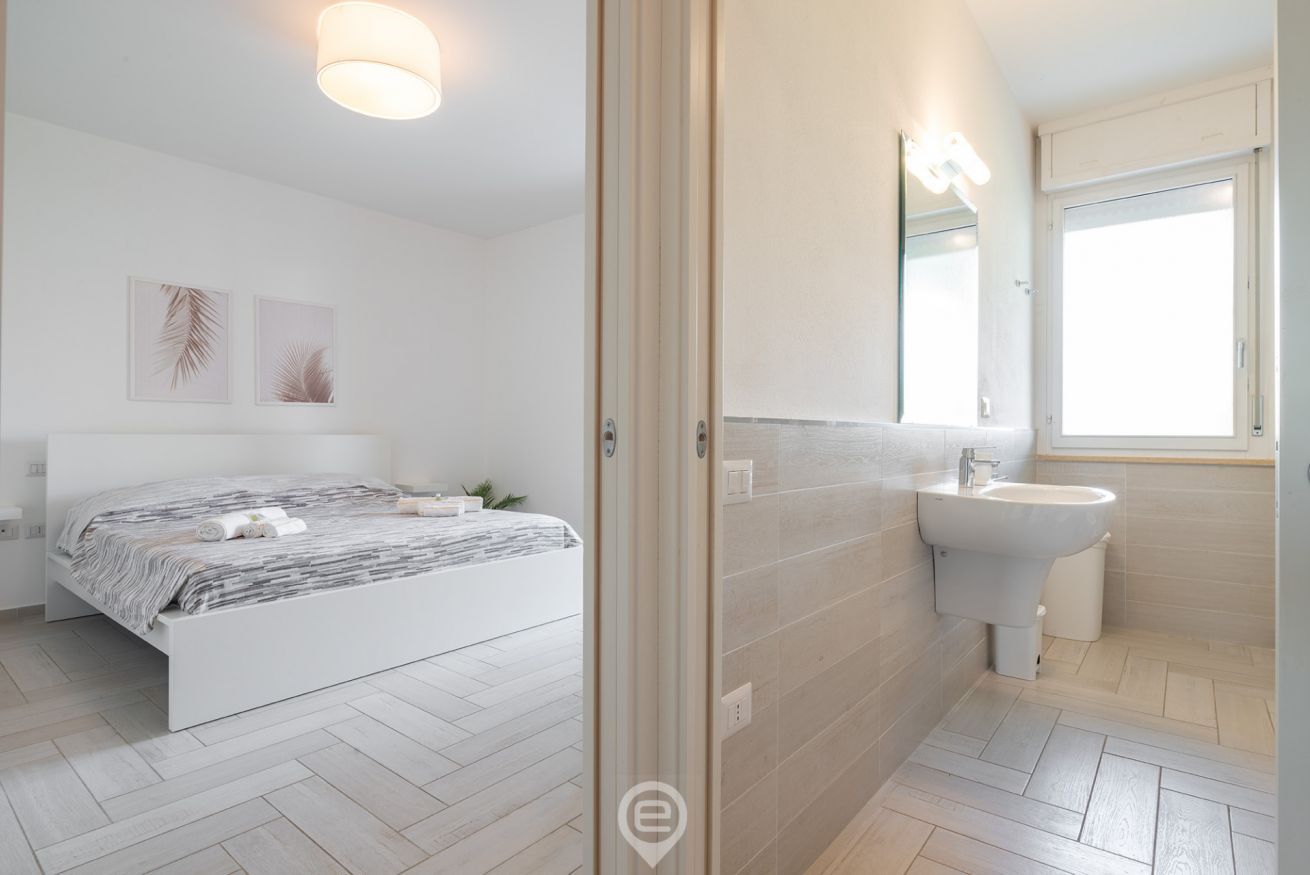 A modern bedroom with a bed, bedside table, and a nightstand. There is a bathroom with a sink and mirror.