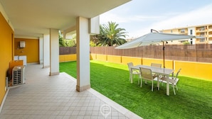 A covered patio with a table and chairs, a small air conditioning unit, and a view of a yellow building and greenery in the background.