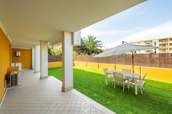 En overdækket terrasse med et bord og stole, en lille aircondition og udsigt til en gul bygning og grøn vegetation i baggrunden.