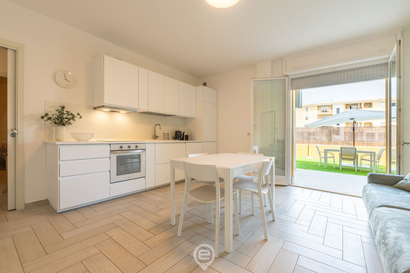 Una cocina moderna con armarios blancos, una mesa de comedor con sillas y vistas a un jardín a través de una puerta corrediza de vidrio.