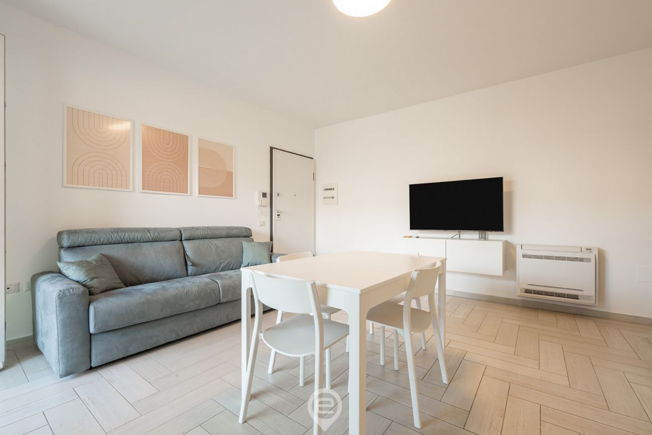 A modern living room with a grey sofa, a white dining table with chairs, a flat-screen TV, and an air conditioning unit.