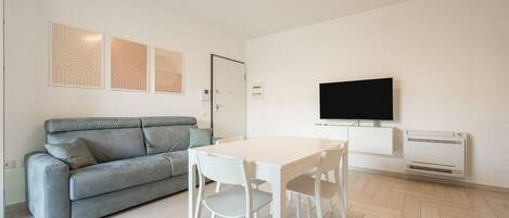 A modern living room with a grey sofa, a white dining table with chairs, a flat-screen TV, and an air conditioning unit.
