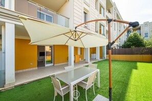 En terrasse med et bord og stole, en udtrækkelig markise, og en bygning med gule vægge og store vinduer.