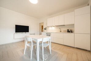 A modern kitchen with white cabinetry, a dining table with chairs, a flat-screen TV, and a built-in oven.