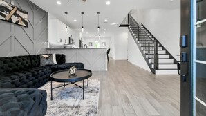 A modern living room with a black sofa, a round coffee table, and a staircase leading to an upper floor. - Grand Luxé Retreat: Lux Home ★ GATED ★ FREE PARKING ★ Medical Center & NRG (Houston)