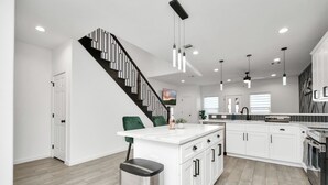 A modern kitchen with white cabinets, a central island, and a black staircase. - Grand Luxé Retreat: Lux Home ★ GATED ★ FREE PARKING ★ Medical Center & NRG (Houston)