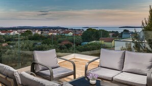A terrace with two grey cushioned chairs and a small table, overlooking a coastal town with houses and greenery.
