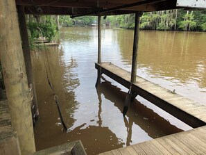 A wooden dock extending over a murky river with trees and a distant structure on the opposite bank.