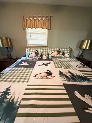 A bedroom with a checkered bedspread featuring bird and tree patterns, a window with blinds, and two bedside lamps. - Two Story House with deck over the river (Karnack)