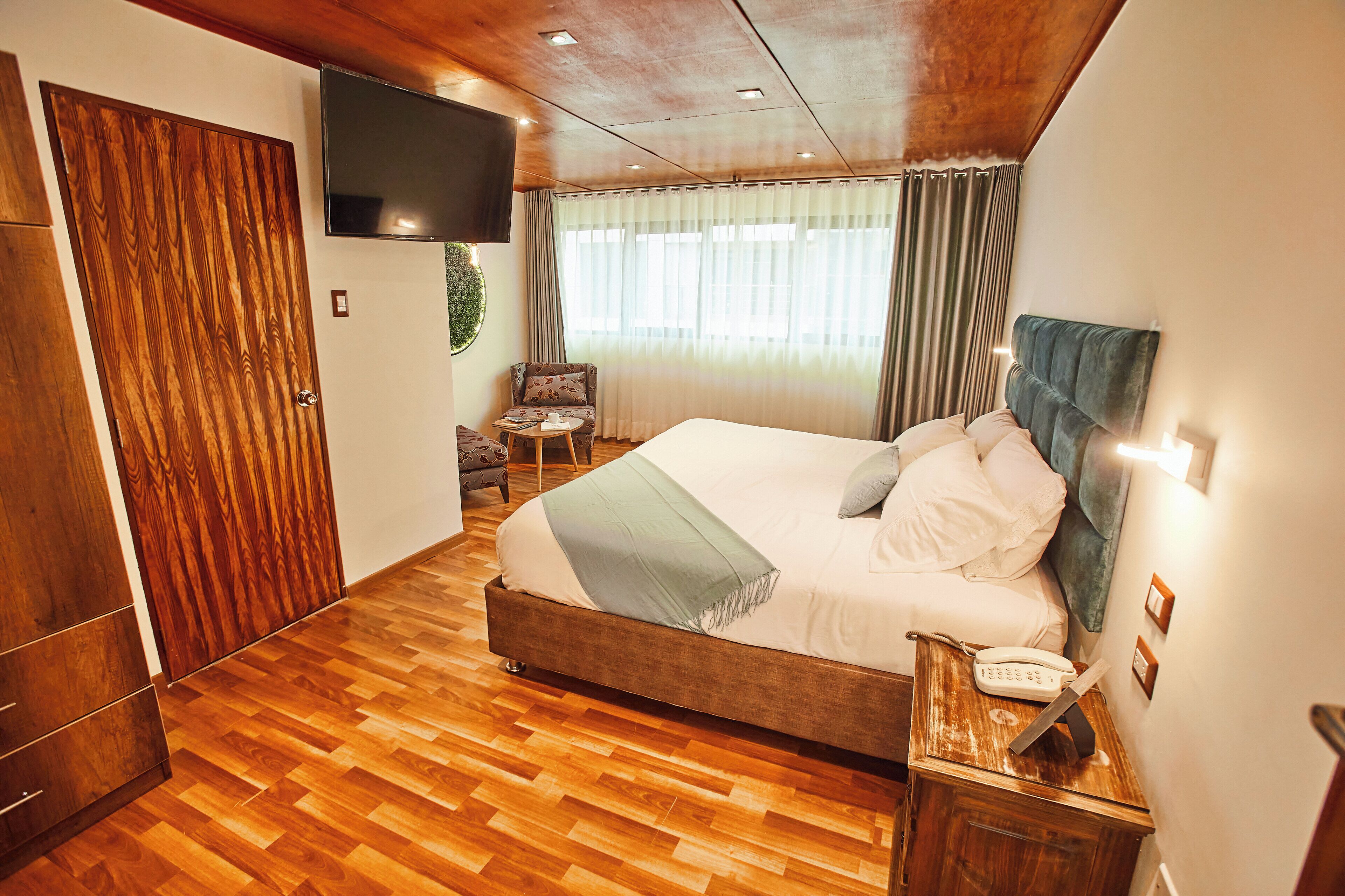 Habitación de hotel con piso de madera, una cama con ropa blanca, una mesita de noche con teléfono, una silla y un televisor en la pared.