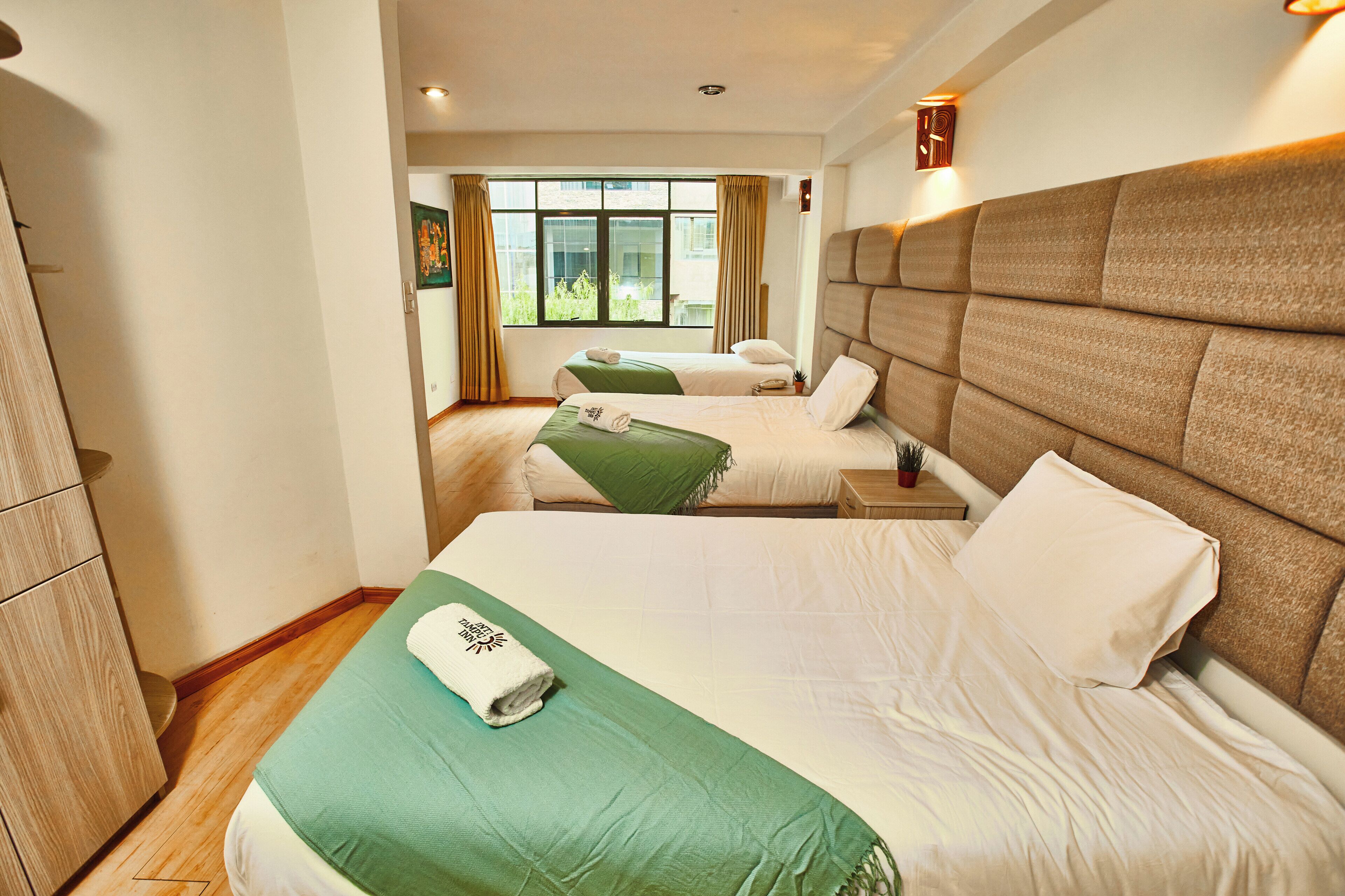 A hotel room with two beds, a wooden wardrobe, a window with curtains, and a wall-mounted light fixture.